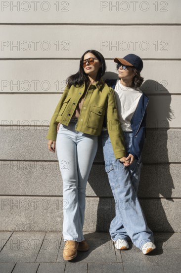 A lesbian couple stands outside, holding hands and enjoying the sunlight. Dressed in casual attire,