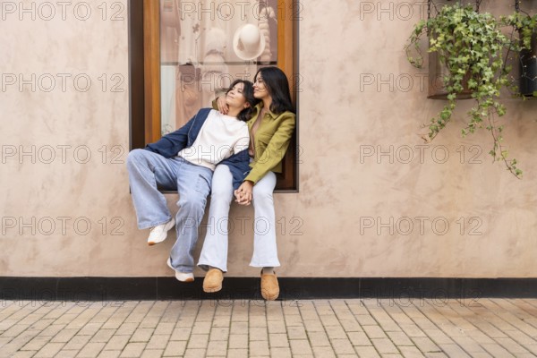 A lesbian couple sits affectionately on a windowsill, holding hands and enjoying a heartfelt moment