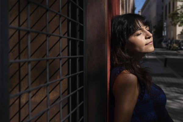A Latin woman leans against a textured wall with her eyes closed, enjoying the warmth of the sun.