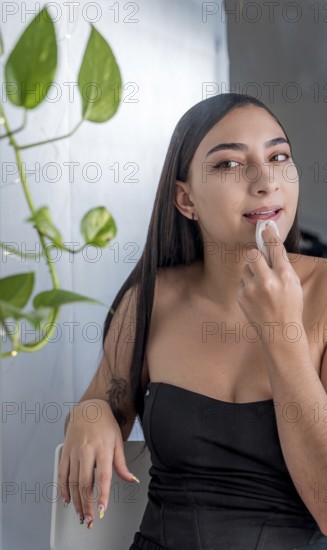 A woman is seated, applying makeup with a sponge in a softly lit room. She wearing a black top,