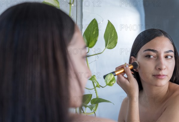 A woman is applying makeup with a brush while looking into a mirror. Green plants add a natural