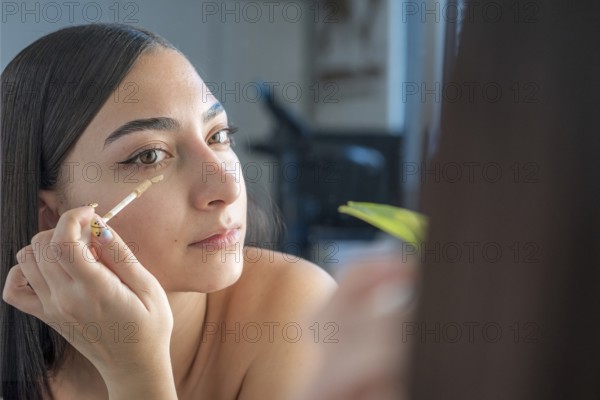 A woman applies concealer under her eyes using a makeup wand, focusing on her reflection in the