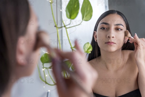 A woman carefully applies makeup using a brush in front of a mirror for the makeup session. Her
