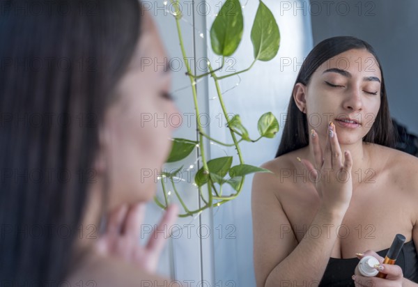 A woman applies skincare cream to her face in front of a mirror. She is focused on self care, with