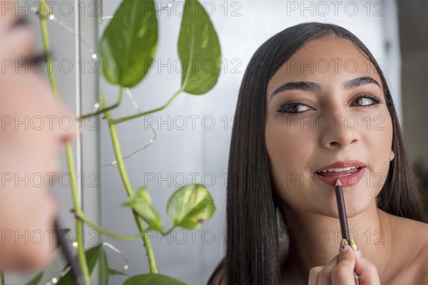 A woman applies lip liner while looking in a mirror, with lush green leaves in the background. The