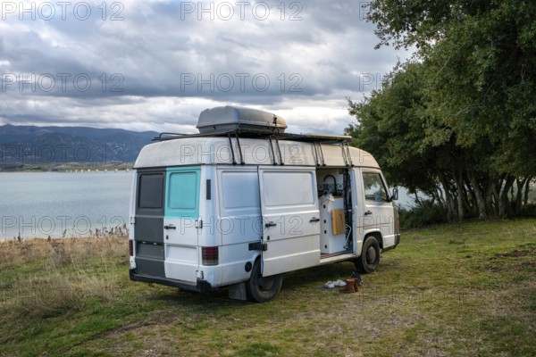 Camper van parked beside a scenic lake in Greece, surrounded by lush greenery and mountains. Cloudy