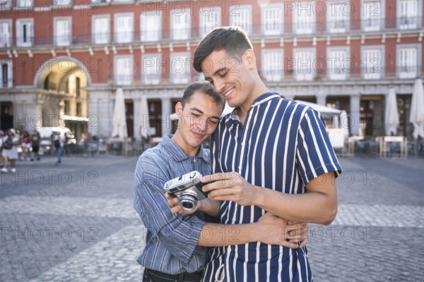 Gay couple embracing while reviewing photos on a vintage camera in Madrid's Plaza Mayor. Warm