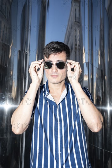 Fashionable young man in a striped shirt adjusting his sunglasses while posing against a reflective