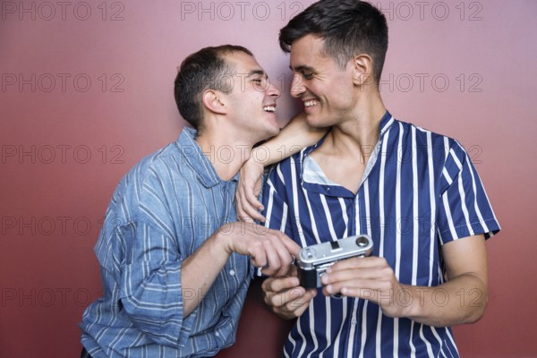 Joyful gay couple sharing a candid moment while holding a vintage camera against a colorful wall.