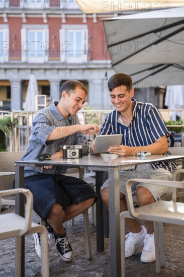 Gay couple sitting at an outdoor cafe in Plaza Mayor, Madrid, smiling while using a tablet and