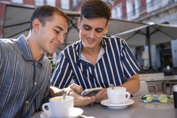 Smiling gay couple enjoying coffee while browsing a smartphone at an outdoor cafe in Plaza Mayor,