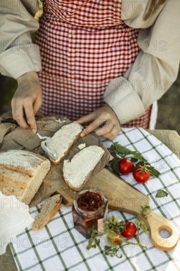 A person wearing a red checkered apron spreads cream on bread slices, surrounded by fresh tomatoes