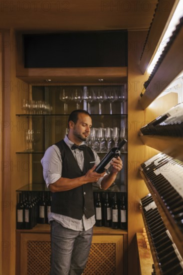 A professional sommelier stands in a cocktail bar, carefully examining a wine bottle. The shelves