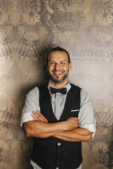 A professional bartender stands confidently with folded arms, wearing a stylish vest and bow tie.