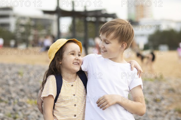 A brother and sister share a joyful moment on a pebble beach. The older boy lovingly wraps his arm