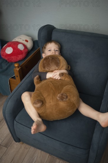 A child relaxes in a cozy chair, cuddling a large plush bear. The scene exudes comfort and warmth,