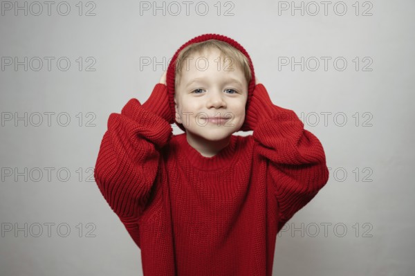 A cheerful young child wearing an oversized red sweater and hat, smiling playfully at the camera.