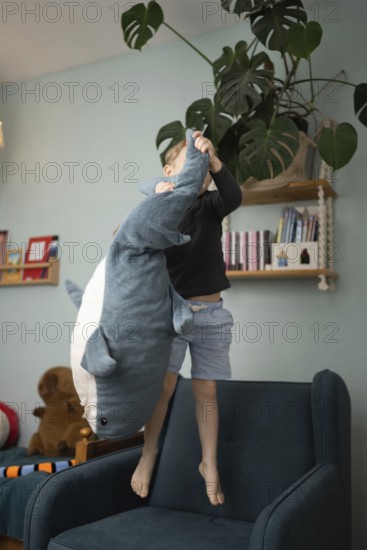A child joyfully playing with a large shark plush toy while standing on a chair in a cozy, well