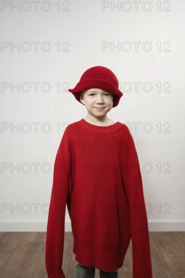 A child wears an oversized red sweater and matching hat, smiling playfully. The neutral background