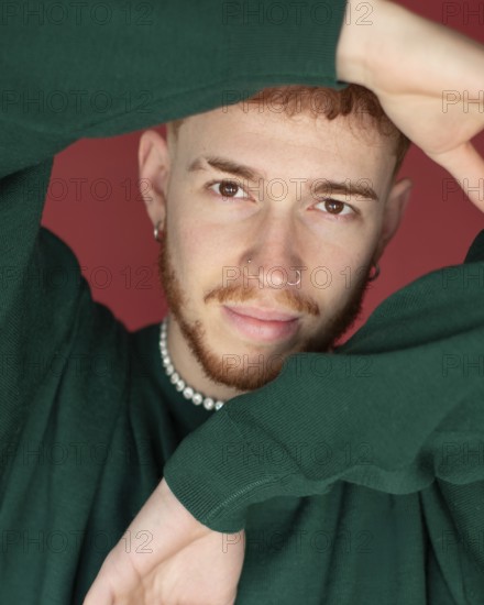 A man red hair wearing a green sweater poses with arms creatively framing their face against a red