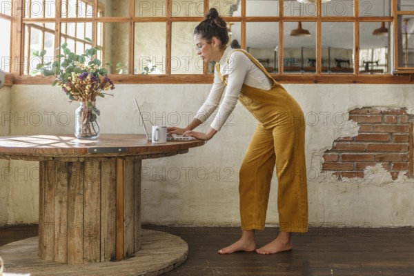 A young woman, dressed in yellow overalls, is focused on her laptop at a wooden circular table in a