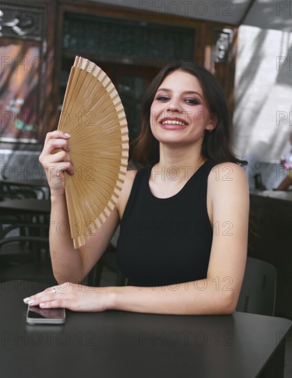 A joyful woman in a black tank top holds a hand fan, sitting at an outdoor cafe table with a