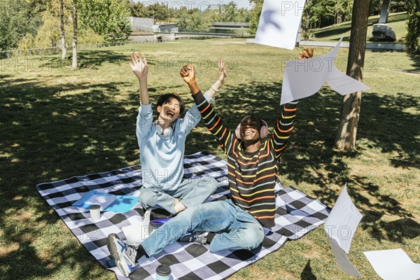 Two university students celebrate joyfully in a sunny park. Sitting on a picnic blanket, they toss