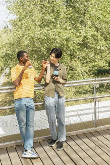 Two friends walking on a deck with lush green trees in the background, enjoying drinks, smiling,