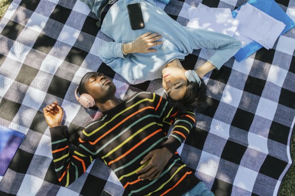 Two friends lie on a checkered blanket outdoors, wearing headphones and enjoying music. Sunlight