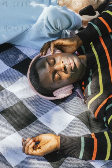 Friends relaxes on a checkered blanket, wearing headphones and basking in the sunlight. The