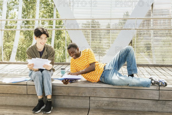 Two friends study outdoors on a sunny day, sitting and laying on a wooden steps surrounded by