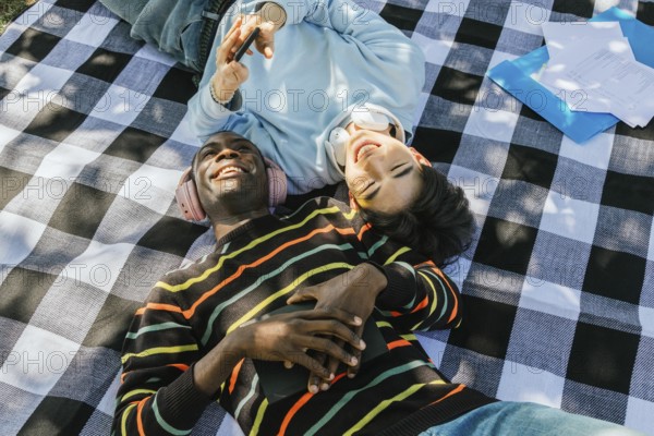 Two friends lie on a checkered picnic blanket, sharing headphones and laughter under the sun. One