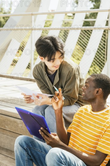 Two friends sit outside on wooden steps, engaged in a study session. One holds a blue folder, and