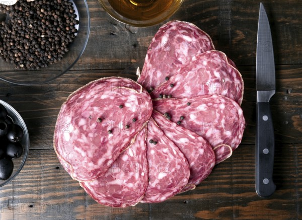 A top down view of sliced spanish salami, peppercorns, black olives, and a knife on a rustic wooden