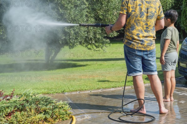 A father and child clean the garden with a pressure washer in summer setting, surrounded by trees