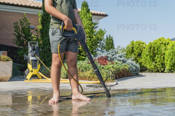 A boy in shorts uses a pressure washer to clean a garden. The washer is in the background, and the