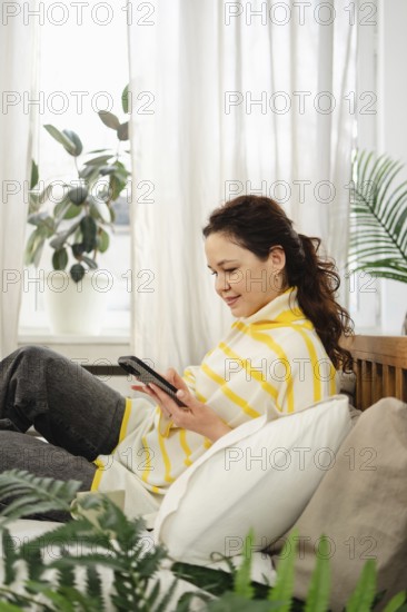 A woman lounges on a sofa, interacting with her smartphone. Surrounded by plush pillows and indoor