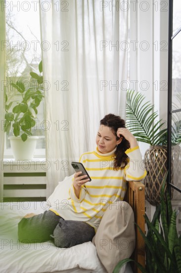 A woman in a striped yellow sweater relaxes on a bed using her smartphone. The room has a cozy