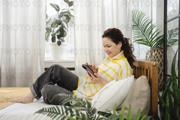 A woman sits comfortably on a bed, wearing a striped top, using her phone. The room is filled with