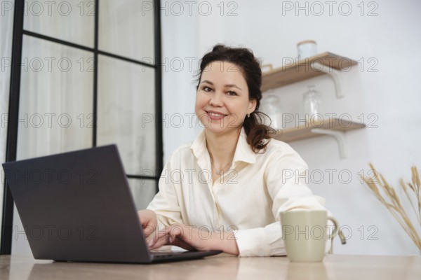 A professional woman, smiling and working on her laptop at a home office desk. Shelves and plants