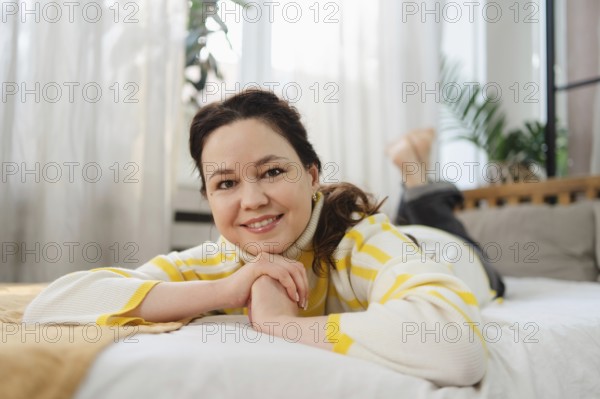 A woman in a yellow striped sweater smiles while lying on a comfortable bed, surrounded by soft