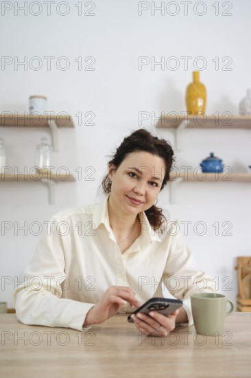 A woman in a white blouse sits at a wooden table in her kitchen, using a smartphone. A mug is