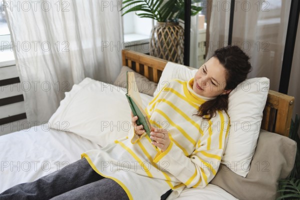 A woman in a yellow striped sweater is relaxing on a bed while reading a book. Soft natural