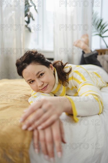 A woman in a striped sweater enjoys a relaxing moment at home. She lies on a bed in a sunlit room,
