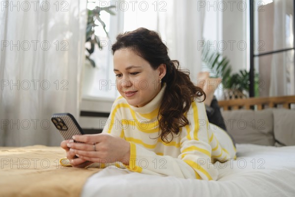 Smiling woman wearing a striped sweater lays on her bed and uses her smartphone. The bright and