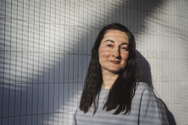 A middle-aged woman smiles gently while standing against a geometrically patterned tile wall, with