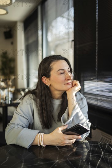A middle-aged woman sits peacefully in a cafe, gazing outside while holding a smartphone in her