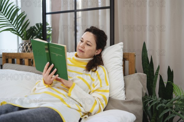 A woman wearing a striped sweater lies on a comfortable bed, deeply engrossed in reading a green