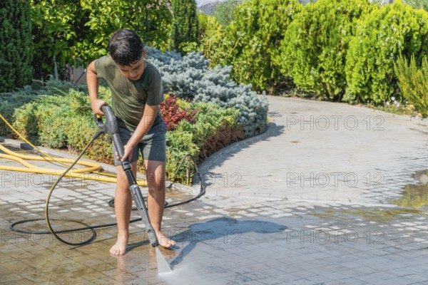 A child is using a pressure washer to clean a stone patio for the summer. He is barefoot, wearing a