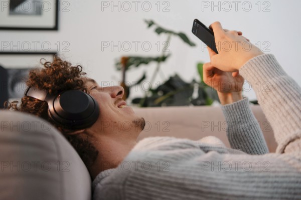A young man with curly hair enjoying leisure time on a sofa, wearing headphones and using a